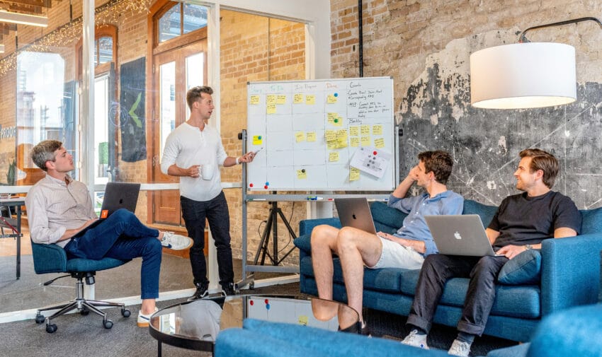 three men sitting while using laptops and watching man beside whiteboard